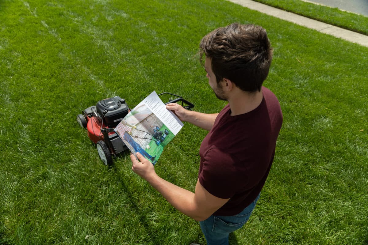 A man reading a manual beside a lawn mower on a grassy area.