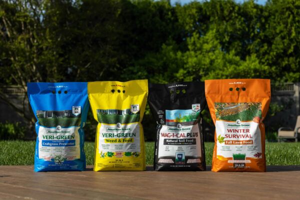 Four bags of Jonathan Green Annual Lawn Care Program for Acidic Soil lined up on a wooden surface, with a vibrant green lawn and trees in the background.