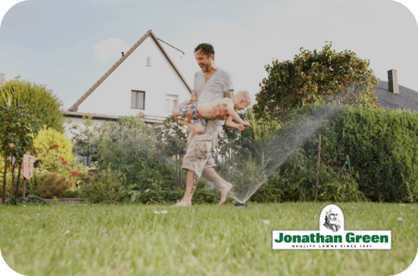Jonathan Green eGift Card runs, carrying a child, through a lawn sprinkler in a backyard.