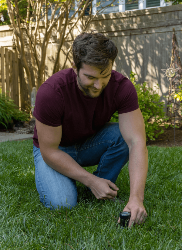Man in a maroon shirt and jeans crouching on a grassy lawn, adjusting a Pro pH & Moisture Soil Tester with his right hand.