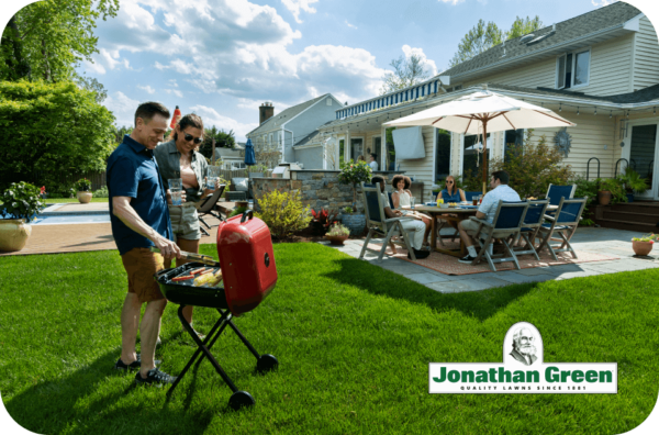 Sentence with replaced product name: Two individuals grilling outdoors while a group of people dine at a patio table in a well-kept backyard, all enjoying the ambiance enhanced by using their Jonathan Green eGift Card for gardening supplies.