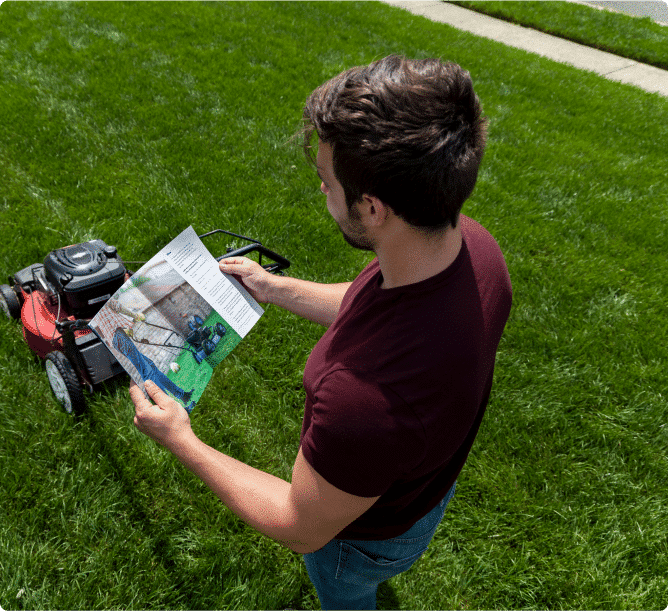 A man reading a manual next to a red lawn mower on a lush green lawn treated with quality grass seed.